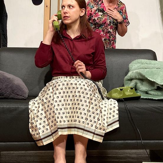 Eine Frau in einem weinroten Hemd und einem gemusterten Rock sitzt auf einer Couch und spricht mit einem grünen Telefon. Eine andere Frau steht hinter ihr und hält ebenfalls ein Telefon.