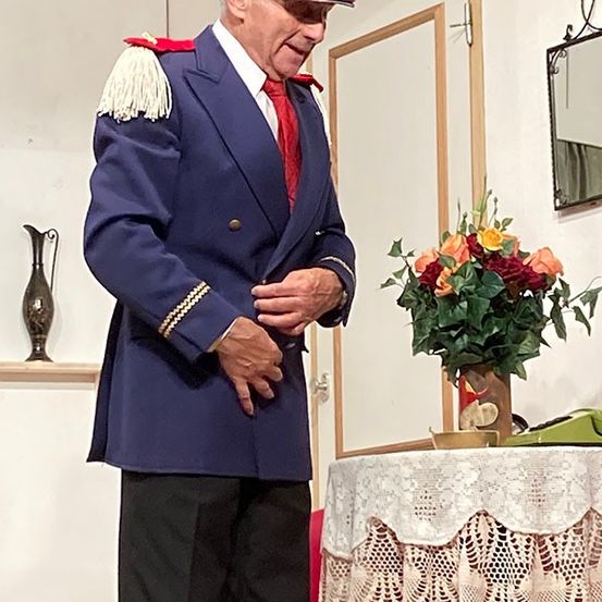 Ein Mann in einem dunkelblauen Uniformmantel mit einer bunten Kappe steht neben einem Tisch mit Blumenarrangement.