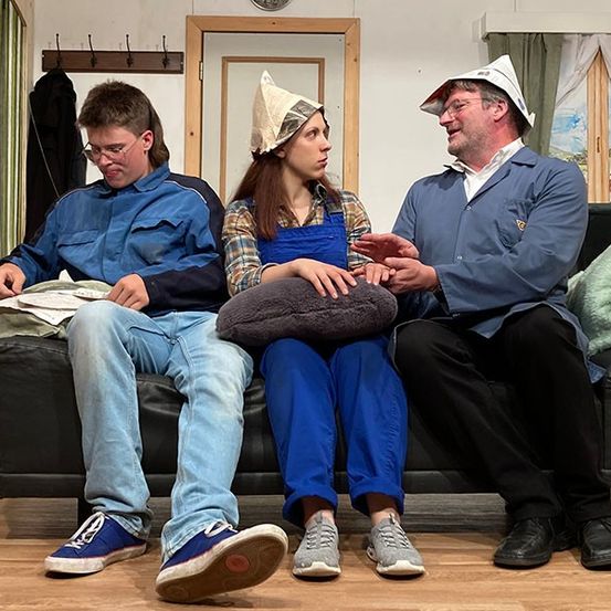 Drei Personen sitzen auf einer Couch in einem Raum und unterhalten sich. Der Mann links trägt eine Brille und eine Jacke. Die Frau in der Mitte hat eine Haube und blaue Latzhosen an. Der Mann rechts, der einen Hut trägt, gestikuliert mit der Hand.