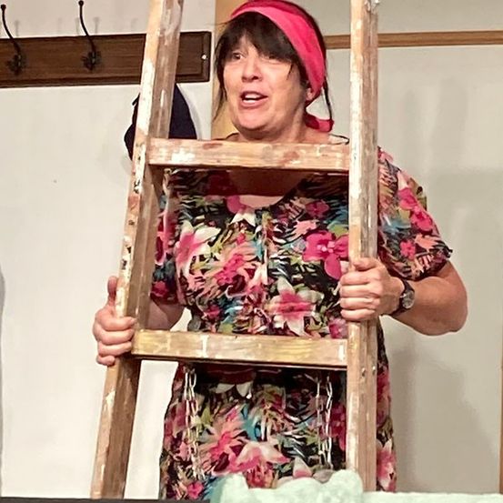 Eine Frau in einem Blumenkleid und einem rosa Kopftuch hält eine hölzerne Leiter und schaut nach oben. Hinter ihr ist eine weiße Wand mit einem hölzernen Kleiderständer zu sehen.