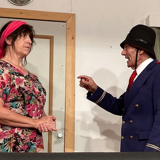 Eine Frau in einem Blumenkleid und einer pinken Kopfbinde steht neben einem Spiegel und sieht einem Mann in einem blauen Uniform und rotem Krawatte zu, der zu schreien scheint.