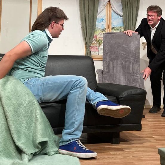 Ein Mann in einem Anzug beugt sich über eine Couch, auf der ein Mann mit Brille sitzt und eine Decke hält. Der Raum hat einen Holzboden und ein Fenster mit einem Gemälde.