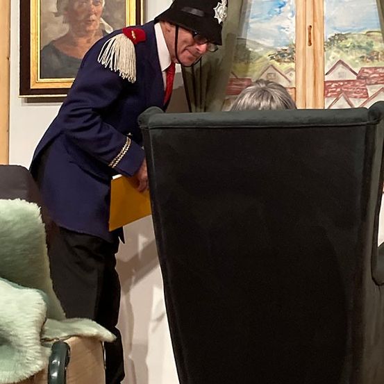 Ein uniformierter Mann schaut unter einen Stuhl und hält eine gelbe Mappe. Er steht in einem Raum mit einem Gemälde an der Wand und einem Fenster mit Blick auf eine Landschaft.