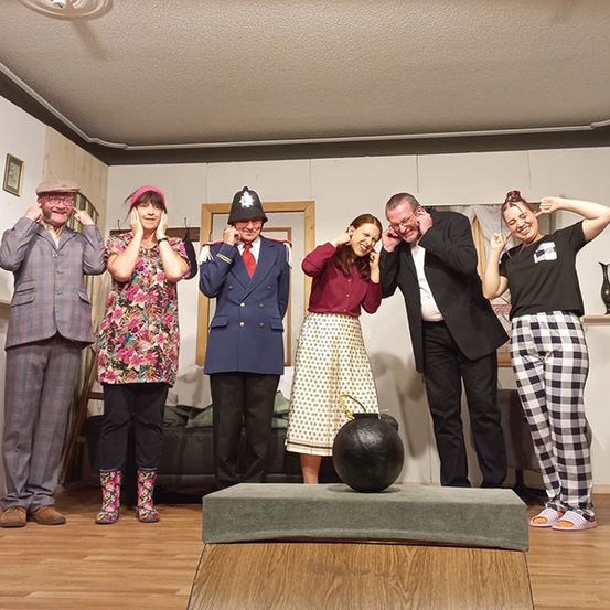 Eine Gruppe Erwachsener in Kostümen posiert auf einer Bühne mit einem großen schwarzen Ball vor ihnen.