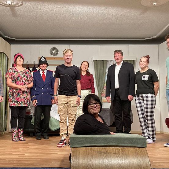 Eine Gruppe von Menschen steht auf einer Bühne, gekleidet in verschiedene Kostüme, wahrscheinlich für eine Aufführung. Eine Frau sitzt in der Mitte auf einem gepolsterten Stuhl.