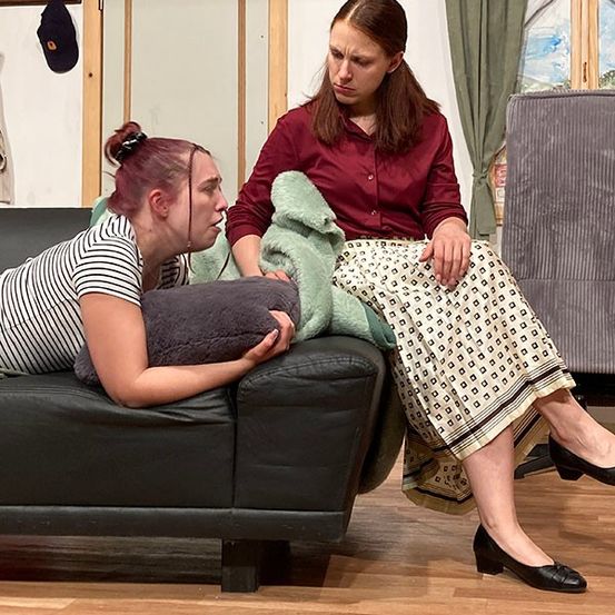 Zwei Frauen sitzen auf einer Couch, eine ruht auf einem Kissen. Die Frau auf der rechten Seite sieht besorgt aus, während die Frau auf der linken Seite verzweifelt wirkt. Hinter ihnen befindet sich eine weiße Tür und grüne Vorhänge.