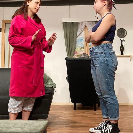 Zwei Frauen stehen sich in einem Wohnzimmer gegenüber, eine in einem rosa Bademantel und die andere in Jeans.