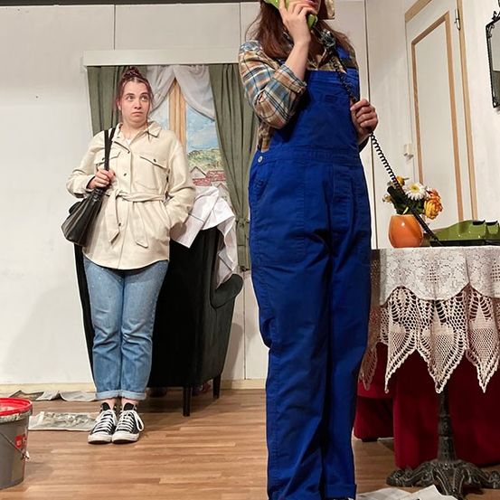 Zwei Frauen in einem Raum, eine am Telefon und die andere mit einer Handtasche. Ein Tisch mit einem Blumenvase steht in der Nähe. Ein Eimer und einige Papiere liegen auf dem Boden.