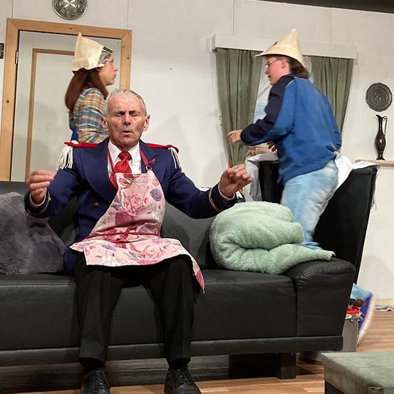 Ein älterer Mann in einem Kostüm sitzt auf einer Couch mit einer Decke, während zwei weitere mit Hüten im Hintergrund stehen.