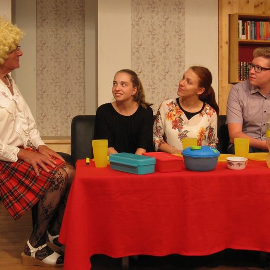 Vier Personen sitzen um einen Tisch herum, darunter eine ältere Frau mit Perücke und drei junge Erwachsene mit Getränken und Essen auf dem Tisch.