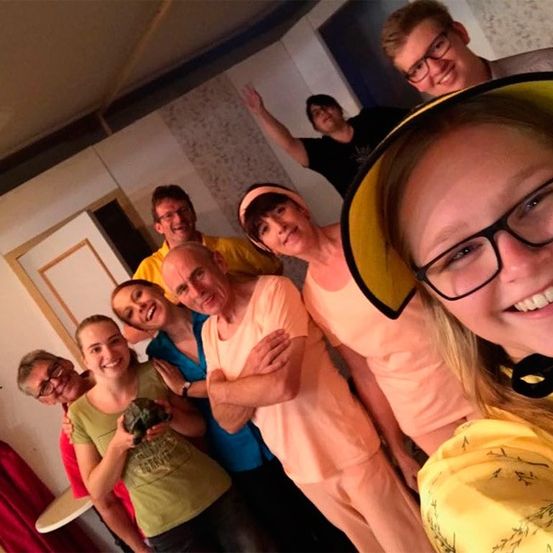 Eine Gruppe lächelnder Personen, einige mit Brille, posiert für ein Foto in einem Raum mit gemusterter Tapete.