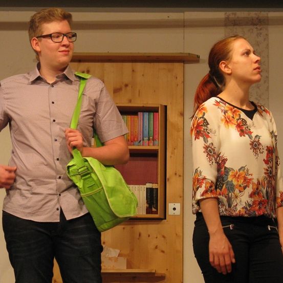 Ein Mann mit Brille und einer grünen Tasche und eine Frau mit einem Pferdeschwanz und einem Blumenhemd stehen neben einem Holzbuchregal.