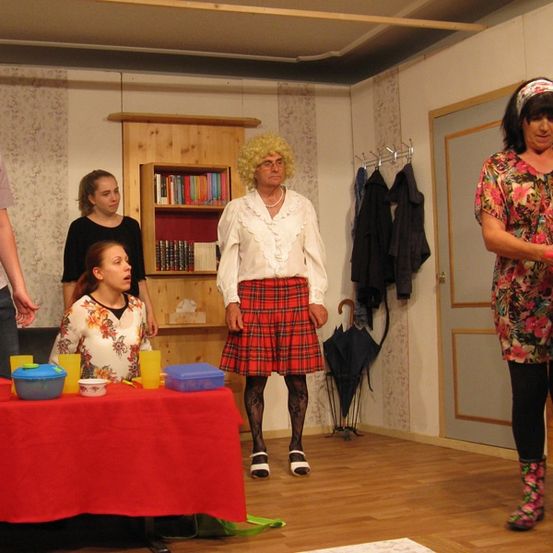 Eine Gruppe von Personen steht um einen Tisch mit verschiedenen Gegenständen. Eine Frau hält einen Blumenstrauß, während andere stehen oder sitzen. Dahinter ist ein Bücherregal und ein Kleiderständer zu sehen.