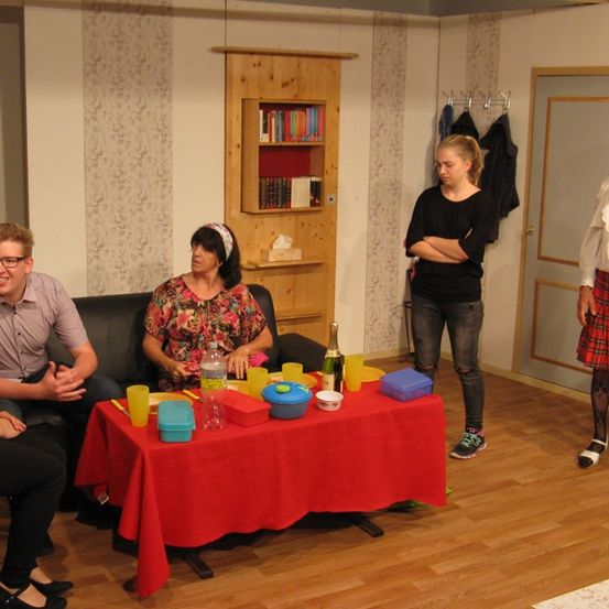 Fünf Personen sitzen um einen Tisch mit roter Tischdecke, auf dem verschiedene Gegenstände wie Tassen und eine Flasche stehen. Eine Frau steht vor ihnen, und eine andere Frau steht in der Nähe einer Tür.