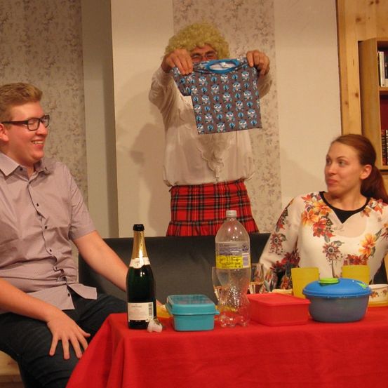 Drei Personen sitzen an einem Tisch, einer hält ein blaues Tuch. Flaschen und Behälter stehen auf dem Tisch. Ein Mann mit Perücke steht hinter ihnen.