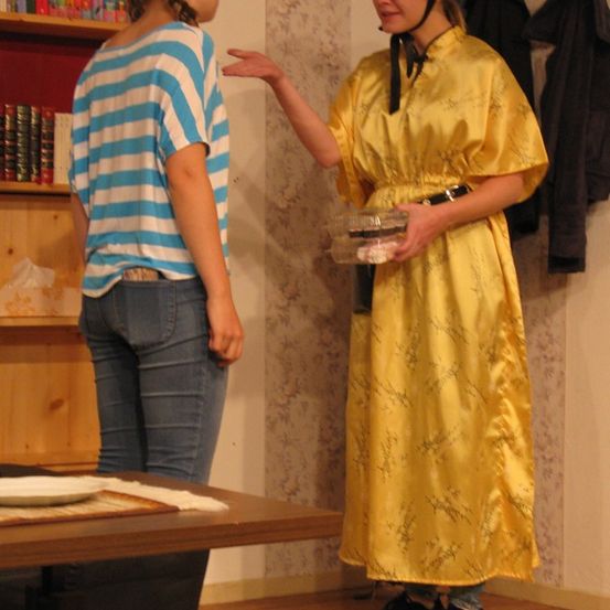 Zwei Frauen unterhalten sich in einem Raum; eine in gelbem Kleid und Hut hält einen Plastikbehälter, während die andere ein gestreiftes Shirt und Jeans trägt. Im Vordergrund steht ein Tisch mit einem Teller.