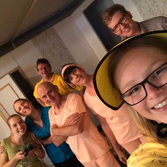 Eine Gruppe von Menschen, wahrscheinlich Schauspieler, lächelt in einem Raum. Sie tragen Brillen und Freizeitkleidung.