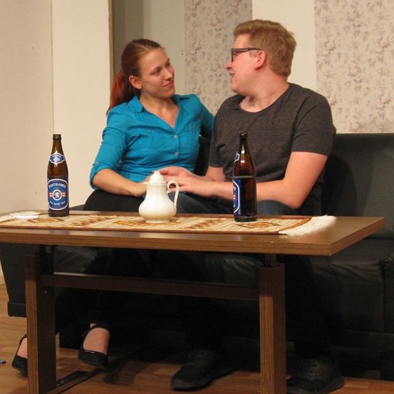 Ein Mann und eine Frau sitzen auf einem Sofa, lächeln und halten Getränke in den Händen, mit einem Tisch davor.