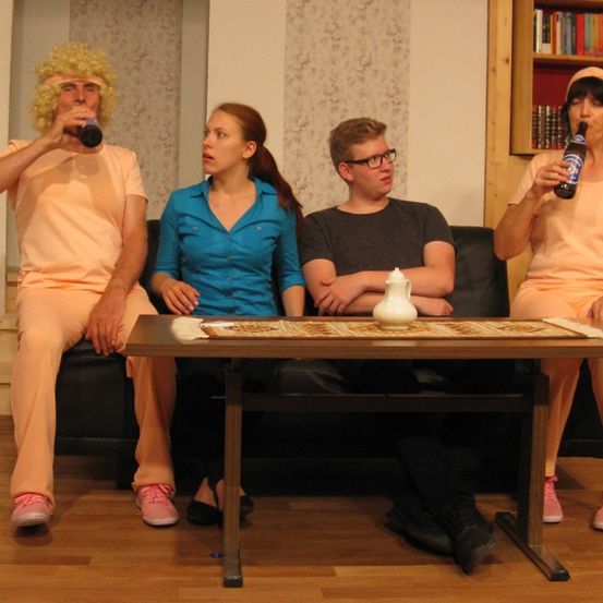 Vier Personen sitzen auf einer schwarzen Couch, alle in passender pfirsichfarbener Kleidung. Sie trinken aus Flaschen. Ein Holztisch steht zwischen ihnen.