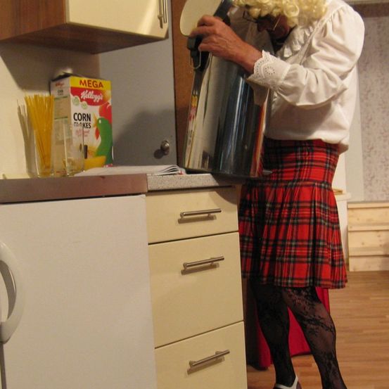Eine Person in einem Kilt und Perücke gießt Kaffee aus einer silbernen Kanne in ein Glas auf einer Küchentheke.