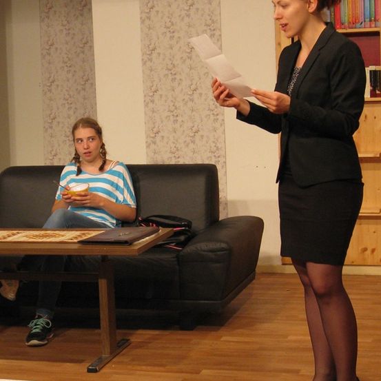 Eine Frau in einem Business-Anzug liest ein Dokument, während sie in einem Wohnzimmer steht, während ein Mädchen in einem gestreiften Shirt auf einem Sofa sitzt, isst und einen Stift in der Hand hält.