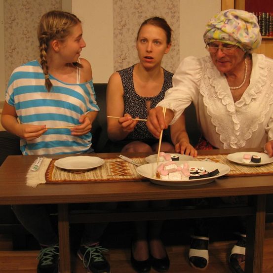 Drei Frauen sitzen auf einer Couch und essen Sushi mit Stäbchen. Eine Frau rechts trägt eine Haube. Ein Tisch vor ihnen hat Teller mit Sushi.