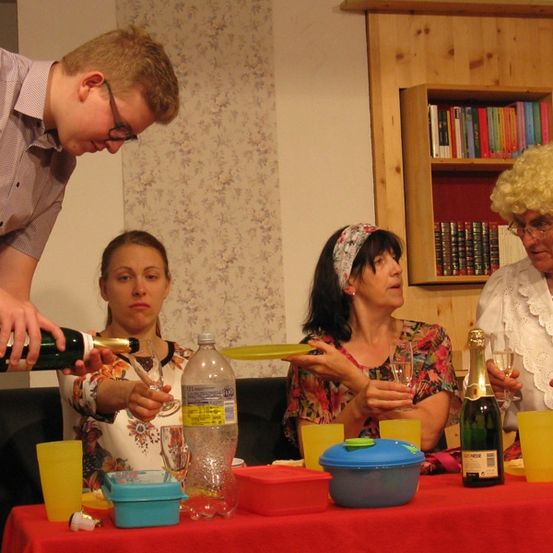 Ein Mann schenkt Wein aus einer Flasche in ein Glas, während drei Frauen um einen Tisch herum sitzen. Eine Frau hält einen Teller, eine andere ein Weinglas und die dritte eine Flasche Champagner.