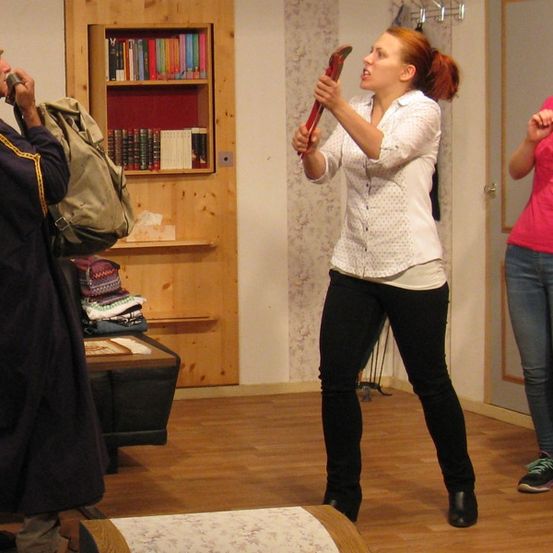 Drei Personen befinden sich in einem Raum mit Holzboden. Ein Mann mit Turban und Robe hält eine Tasche, eine Frau in einem weißen Hemd hält ein Werkzeug und eine andere Frau in einem rosa Shirt steht in der Nähe.