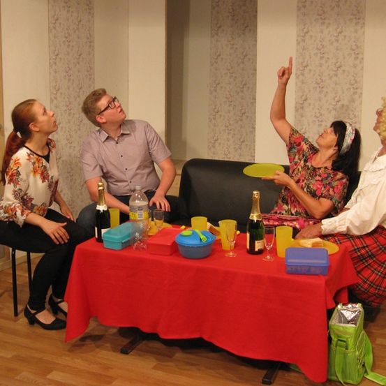 Vier Personen versammeln sich um einen Tisch, eine zeigt mit dem Finger, eine hält einen gelben Teller und zwei sitzen, mit Flaschen und Gläsern auf dem Tisch.