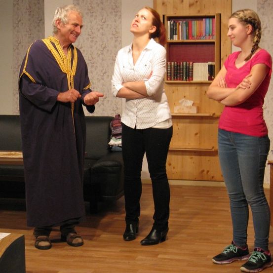 Drei Menschen stehen in einem Raum. Ein Mann in einem Gewand und Sandalen, eine Frau in einem weißen Hemd und eine weitere Frau in einem rosa Shirt. Die Frau im weißen Hemd schaut nach oben, während der Mann lächelt.