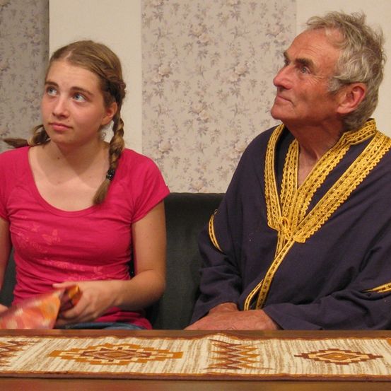 Ein älterer Mann und ein junges Mädchen sitzen auf einer Couch, wobei das Mädchen ein Stück Stoff hält und den Mann ansieht, der einen violetten Umhang mit goldener Einfassung trägt.