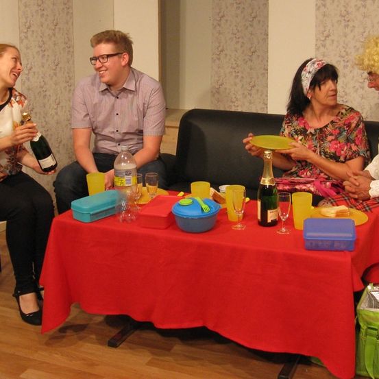 Vier Personen sitzen um einen Tisch herum, mit jeweils zwei Frauen auf beiden Seiten. Eine Frau hält eine Flasche, während die andere einen gelben Teller hat. Ein Mann sitzt zwischen ihnen, und der Tisch ist mit Gläsern, einer Flasche und verschiedenen Gegenständen gedeckt.