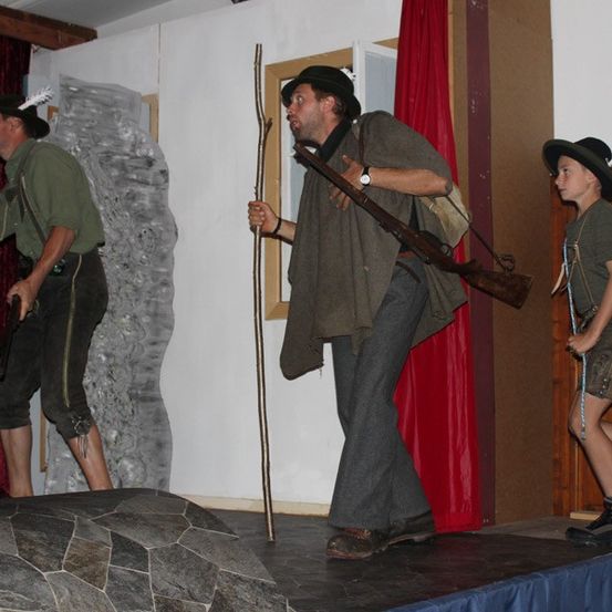 Drei Männer stehen auf der Bühne, zwei halten Gewehre und einer eine lange Stange. Sie tragen Hüte und grüne Hemden. Hinter ihnen ist eine Kulisse aus roten Vorhängen und einer Steinmauer.