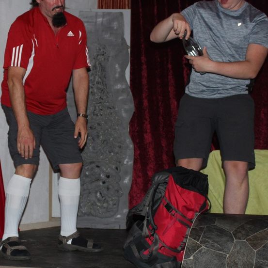 Zwei Männer stehen auf einer Bühne, einer hält eine Glasflasche und der andere einen Rucksack. Im Hintergrund befinden sich ein Vorhang und eine Steinstatue.