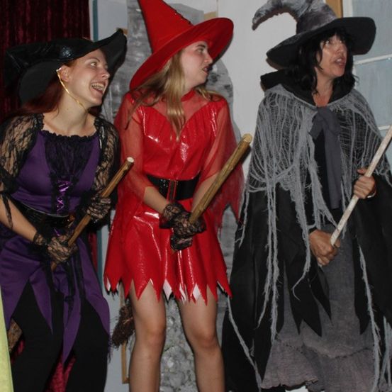 Drei Frauen in Hexenkostümen mit roten Hüten und Besen stehen zusammen, lächeln und posieren für ein Foto.