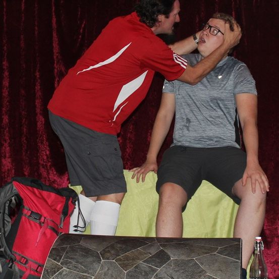 Ein Mann in einem roten Shirt justiert die Brille eines anderen Mannes in einem grauen Shirt auf der Bühne, während eine rote Tasche und eine Flasche in der Nähe sind.