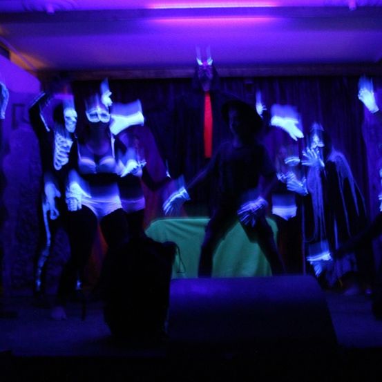 Eine Gruppe von Menschen in Kostümen tritt auf der Bühne bei lila Beleuchtung auf. Sie haben leuchtende Elemente an Körper und Händen.