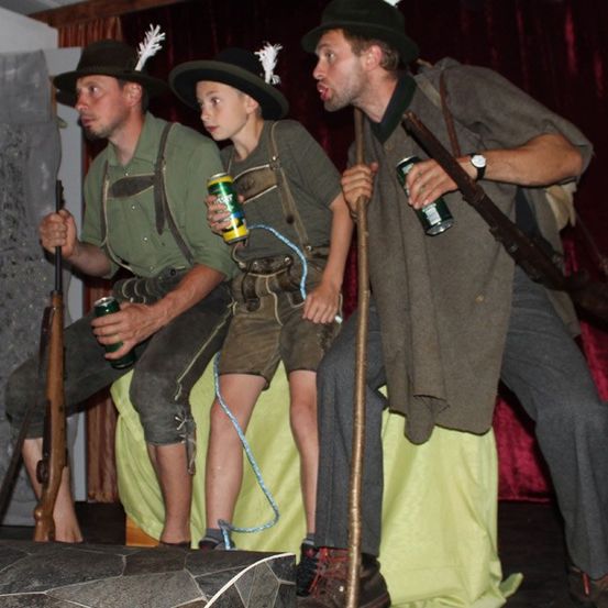 Drei Männer, gekleidet in traditioneller Kleidung, stehen auf einer Bühne und halten Gewehre. Einer hält eine Bierdose, während die anderen beiden Stöcke halten. Sie scheinen in einem Theaterstück aufzutreten.