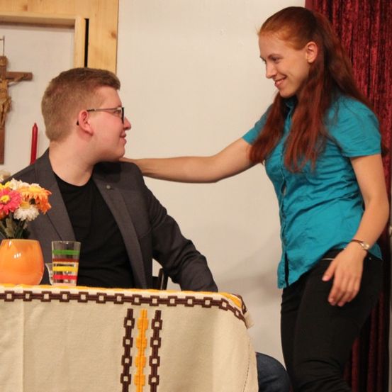 Ein junger Mann und eine junge Frau sitzen an einem Tisch, wobei die Frau sich über den Mann beugt, während er lächelt. Auf dem Tisch steht eine Vase mit Blumen und ein Glas Wasser. Hinter ihnen befindet sich ein Kreuz und rote Kerzen.