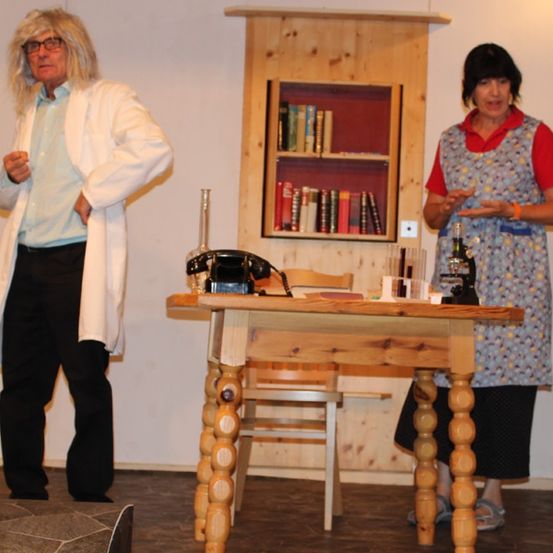 Zwei Personen auf der Bühne, eine in einem Laborkittel und die andere in einem Kleid, stehen neben einem Holztisch und einem Regal mit Büchern.