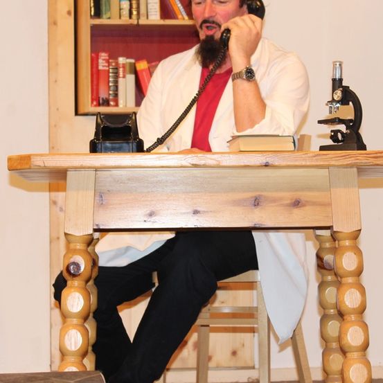 Ein Mann in einem weißen Laborkittel sitzt an einem Holzschreibtisch und hält ein Telefon. Bücher, ein Mikroskop und eine Uhr liegen auf dem Tisch. Dahinter steht ein hölzerne Bücherregal, gefüllt mit Büchern.