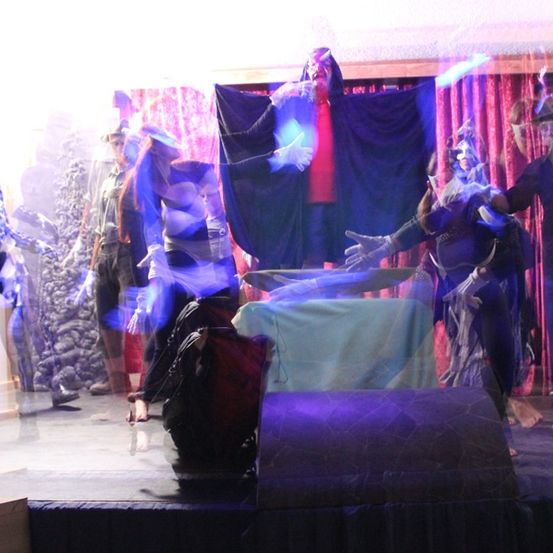 Eine Bühnenaufführung mit Menschen in Kostümen und Lichteffekten, eine Skelettfigur links und eine Gruppe in der Mitte.