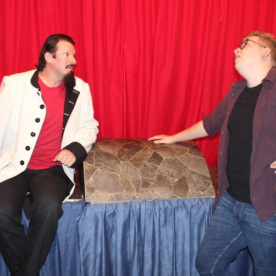 Zwei Männer sitzen auf einem Tisch mit einem roten Vorhang im Hintergrund. Einer trägt einen weißen Mantel und ein rotes Shirt, der andere ein kariertes Hemd und eine Brille.