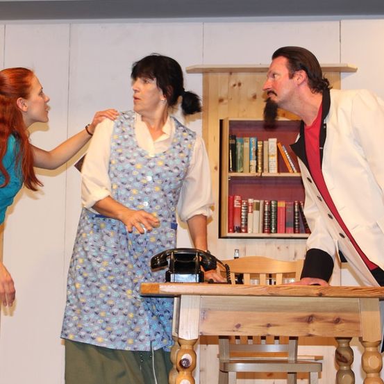 Drei Personen stehen um einen Tisch herum, wahrscheinlich in einer inszenierten Szene. Eine Frau mit roten Haaren lehnt sich vor, während eine Frau in einem Kleid zu einem Mann in einem weißen Mantel spricht.