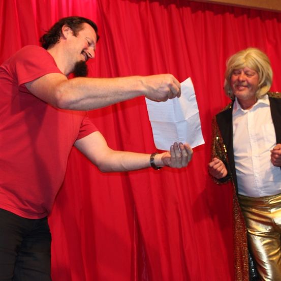 Ein Mann in einem roten T-Shirt und schwarzen Hosen hält ein Stück Papier und gibt es einem anderen Mann in einem goldglänzenden Jackett auf der Bühne.