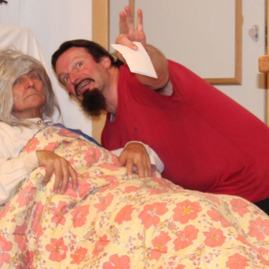 Ein Mann in einem roten T-Shirt hält ein weißes Objekt, während eine ältere Frau mit grauem Haar in einem Bett mit einem Blumenlaken liegt.