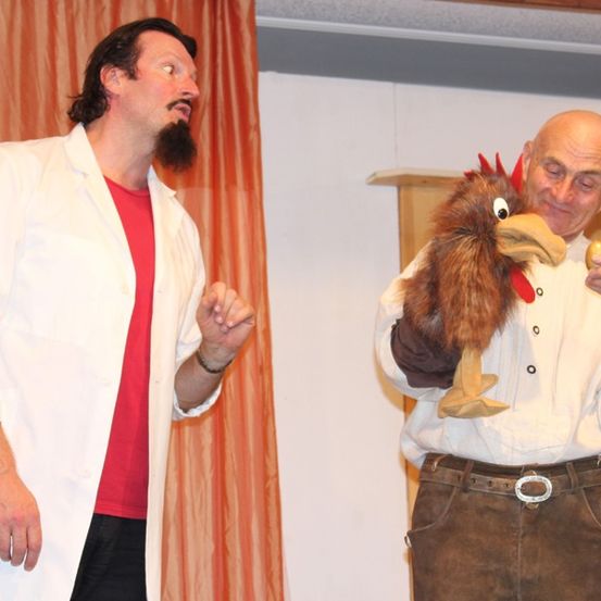 Zwei Männer auf einer Bühne, einer in einem weißen Mantel und der andere in einer braunen Weste, führen ein Theaterstück auf.