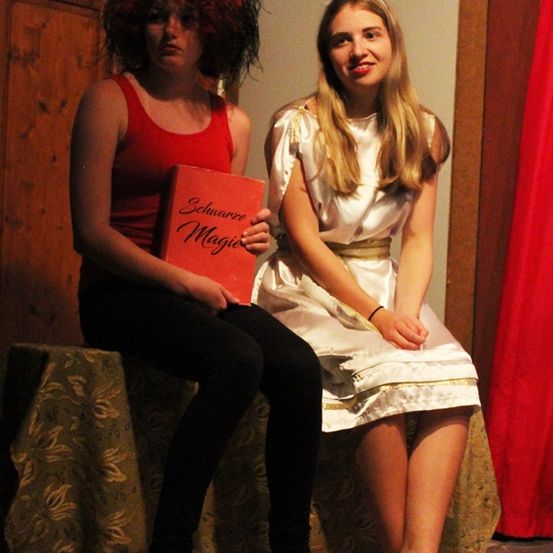 Zwei Mädchen sitzen auf einer Bank. Eine hält ein Buch, die andere trägt einen Heiligenschein. Sie scheinen auf einer Bühne zu sein.