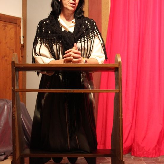 Eine Frau in Schwarz und Weiß steht hinter einem hölzernen Podium und schaut auf einen roten Vorhang.