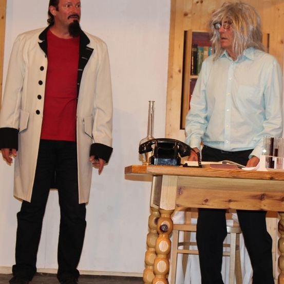 Zwei Männer auf der Bühne, einer in einem weißen Mantel mit Bart, der andere in einem blauen Hemd mit Brille, stehen neben einem Holzschreibtisch mit einem Telefon.